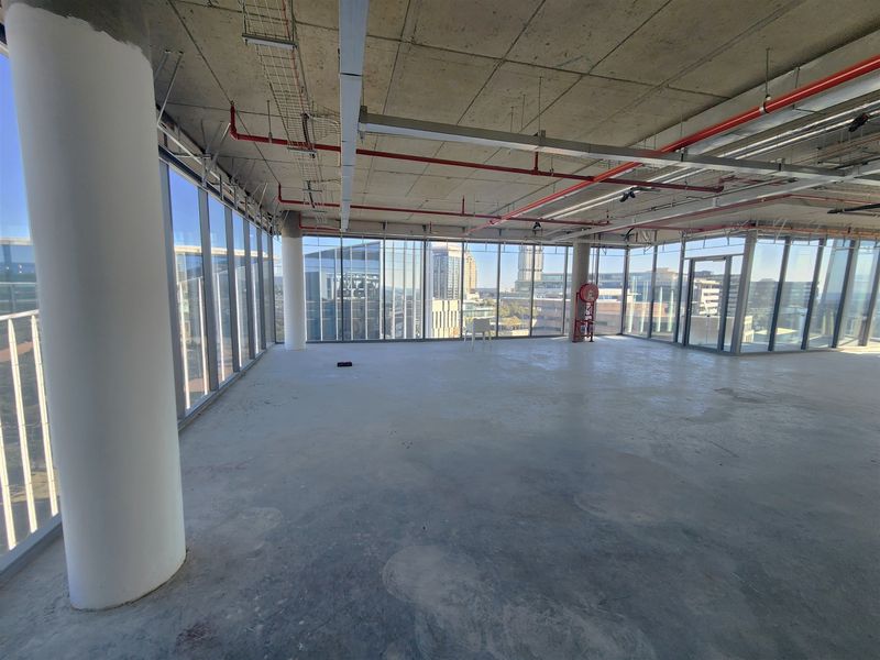 An expansive, open-plan office space in shell condition, featuring floor-to-ceiling windows with city views and exposed industrial-style ceiling services.