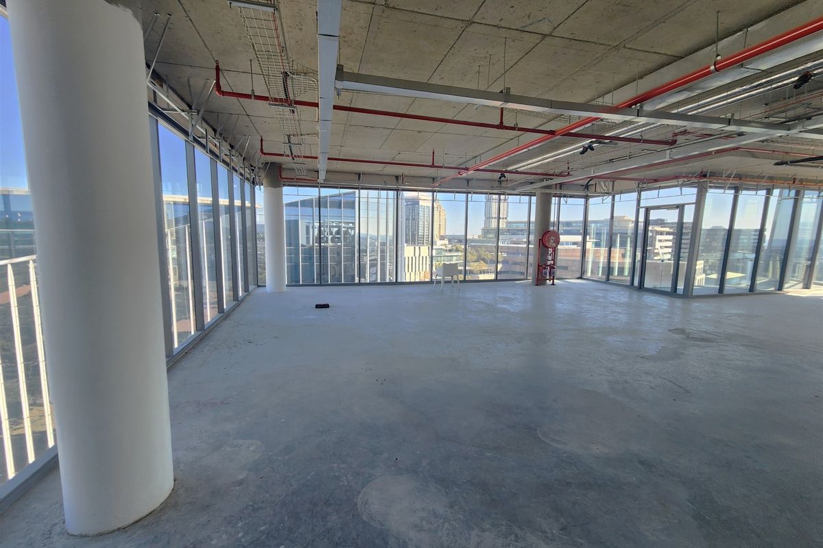 An expansive, open-plan office space in shell condition, featuring floor-to-ceiling windows with city views and exposed industrial-style ceiling services.