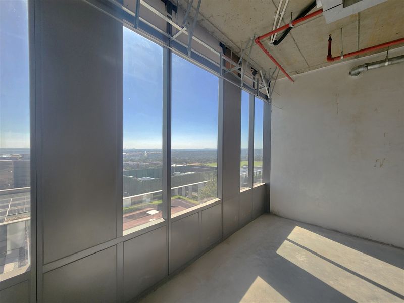 An unfinished office space featuring floor-to-ceiling windows with expansive views, exposed concrete ceilings, and industrial fire sprinkler systems.