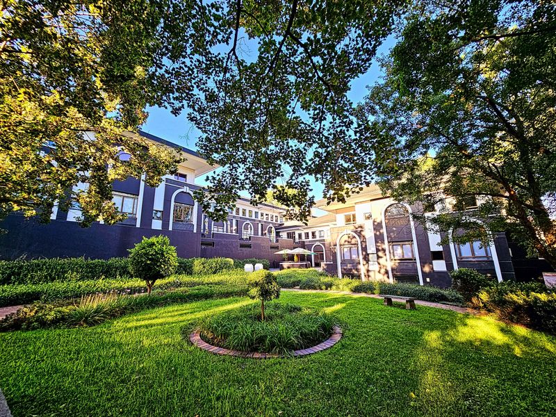 A well-maintained rear garden featuring manicured lawns, brick-edged garden beds, and a stone bench, set against the backdrop of a multi-story office building.