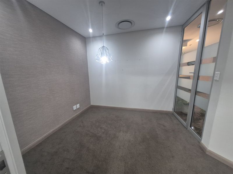 A clean, empty office space featuring neutral textured wallpaper, carpet flooring, a modern pendant light, and a glass partition door.