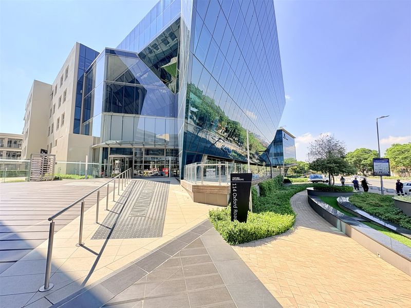 Modern commercial office building exterior featuring a striking glass facade, paved pedestrian entrance, and well-maintained landscaped grounds.