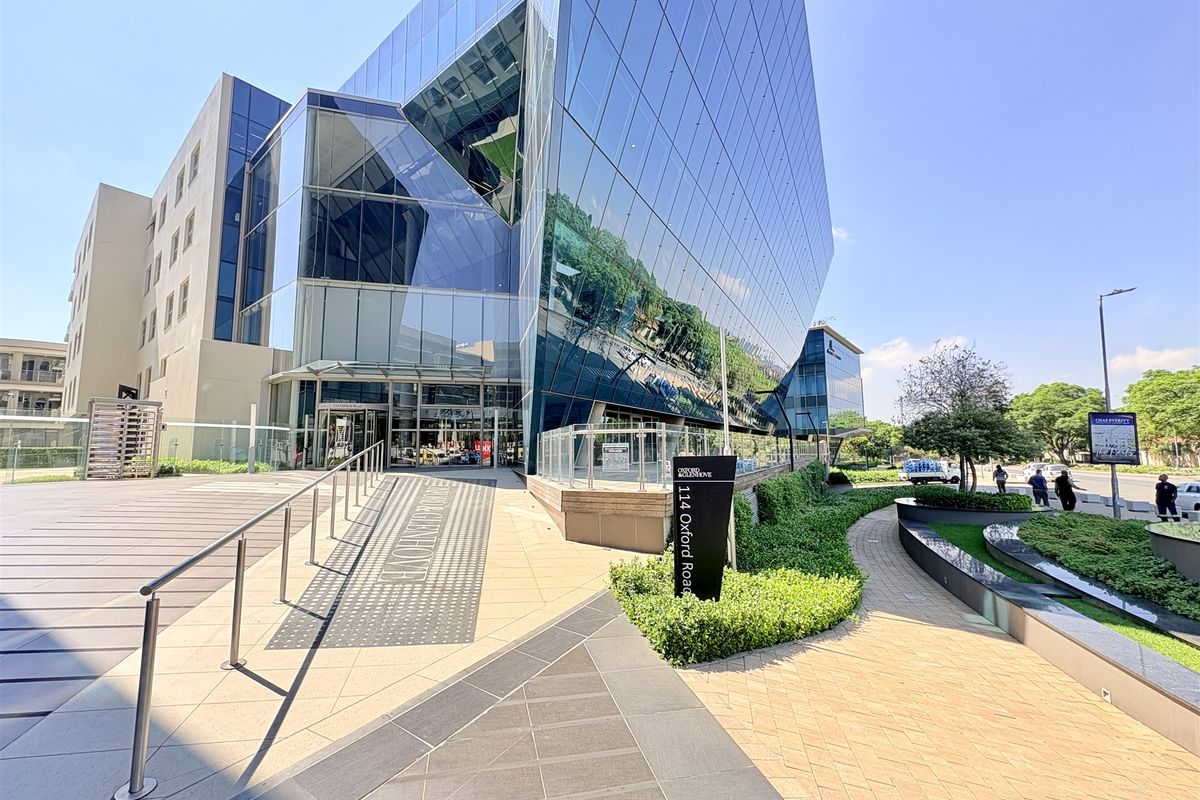 Modern commercial office building exterior featuring a striking glass facade, paved pedestrian entrance, and well-maintained landscaped grounds.