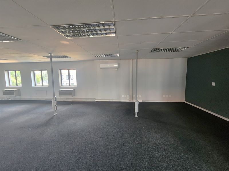 An empty open-plan office space featuring suspended ceilings, recessed lighting, wall-mounted air conditioning units, and carpeted flooring.