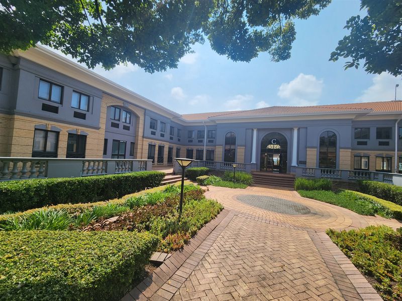A well-maintained office building exterior featuring a paved courtyard, landscaped gardens, and a prominent arched entrance.