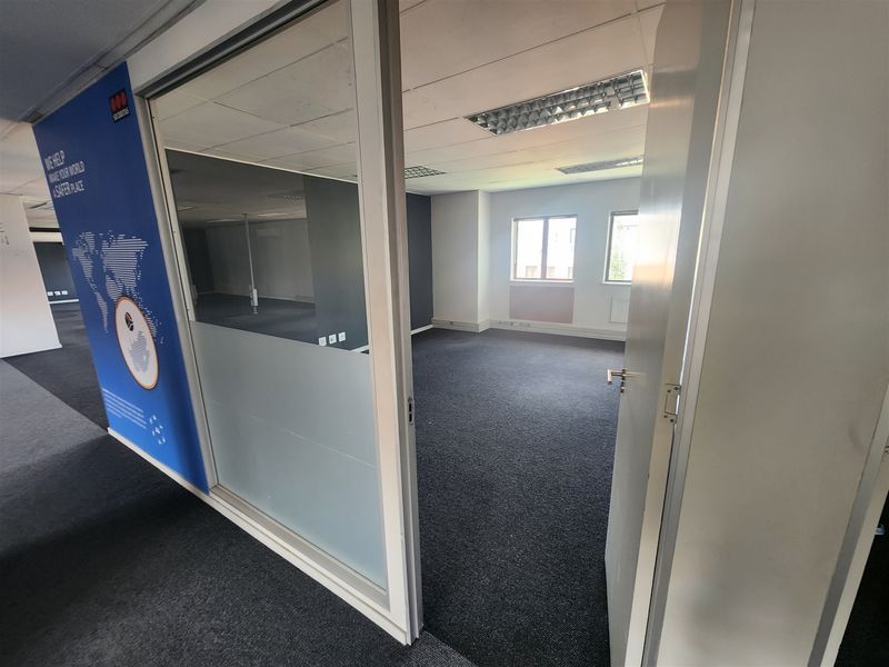 An empty office space featuring glass partitioning, carpeted flooring, and a suspended ceiling with recessed lighting, offering a clean and functional workspace environment.