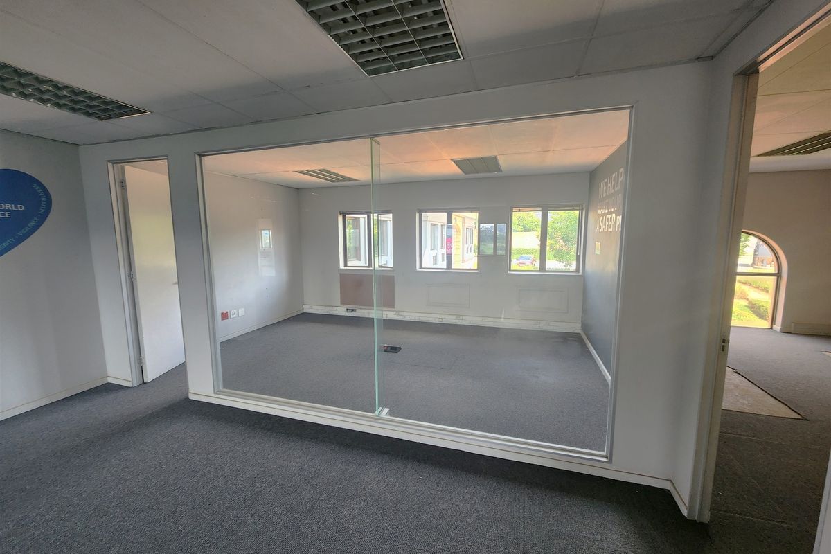 An empty office space featuring a large glass partition wall, suspended ceiling with recessed lighting, and carpeted flooring.