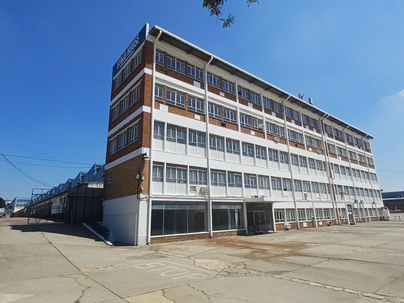 A multi-story industrial building featuring a brick and white panel facade with extensive window glazing, situated on a large paved industrial site.
