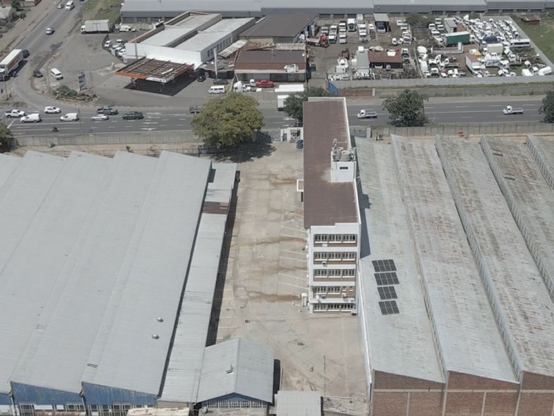 Aerial view of an industrial property featuring large warehouse structures, a multi-story office block, and a paved yard area with solar panels on the roof.