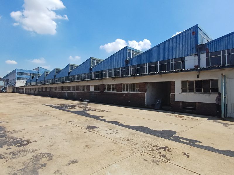 Exterior view of an industrial warehouse facility featuring a large concrete yard and sawtooth roof design with clerestory windows.