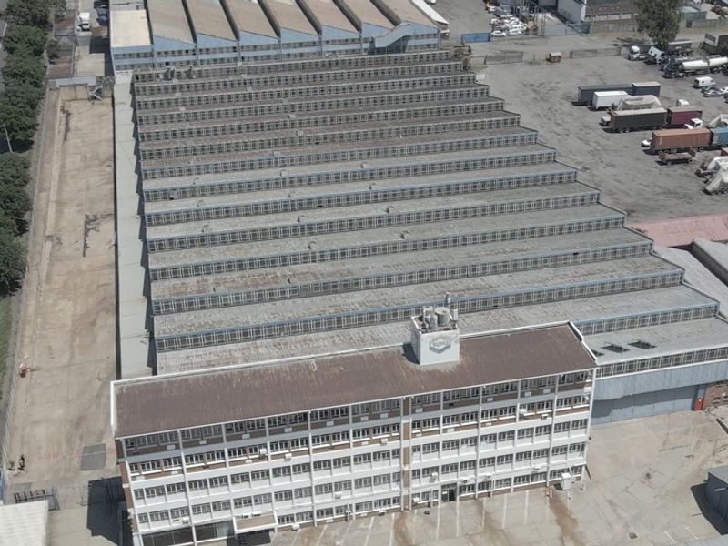 Aerial view of a large industrial facility featuring a multi-story office block attached to an expansive warehouse with sawtooth roofing.