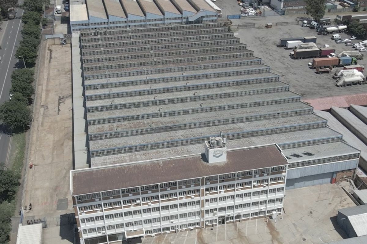 Aerial view of a large industrial facility featuring a multi-story office block attached to an expansive warehouse with sawtooth roofing.
