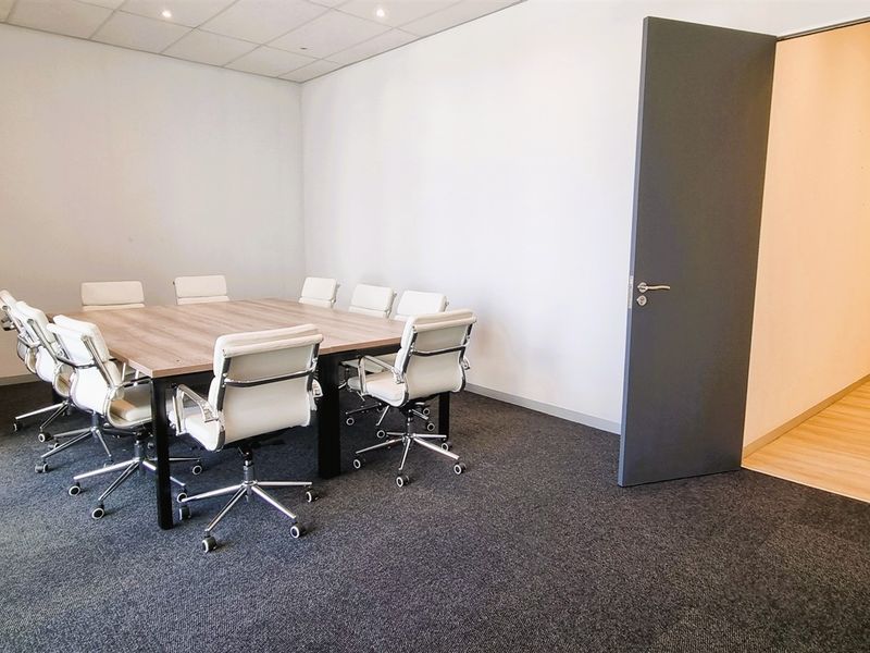 A professional office meeting room featuring carpeted flooring, a suspended ceiling with recessed lighting, and direct access to a hallway.