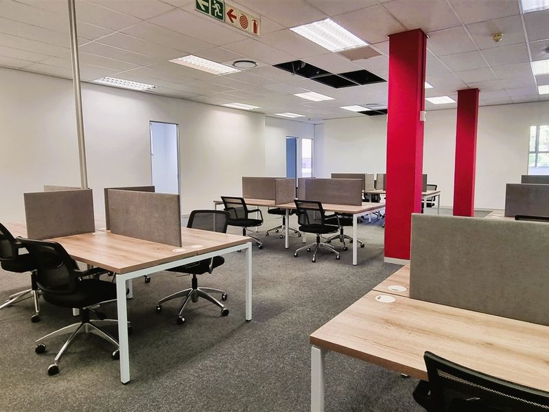 Open-plan office space featuring carpeted flooring, suspended ceiling with recessed lighting, and prominent structural columns.
