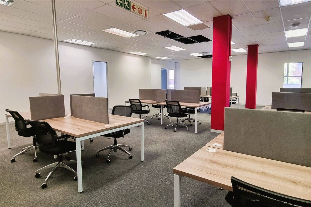 Open-plan office space featuring carpeted flooring, suspended ceiling with recessed lighting, and prominent structural columns.