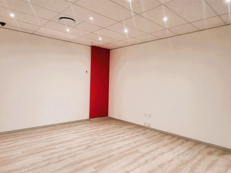 A clean, empty office space featuring modern laminate flooring, a suspended ceiling with recessed lighting, and a distinctive red accent wall.