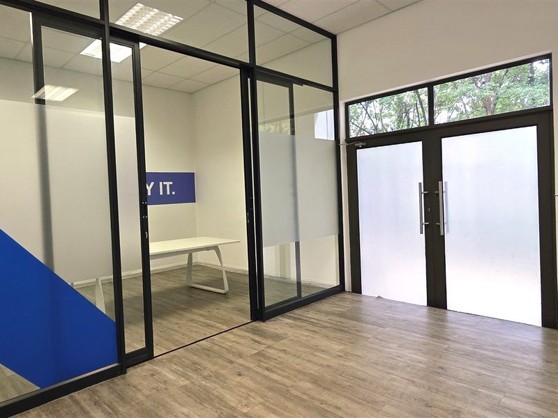 Modern office space featuring glass partition walls, laminate flooring, and a bright, open layout with double glass doors.