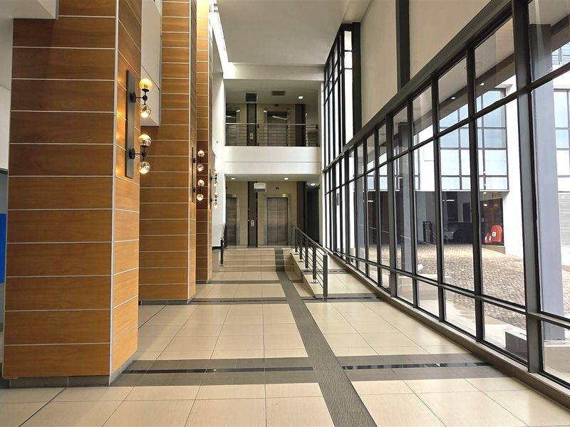 Spacious modern office building lobby featuring floor-to-ceiling glass walls, wood-panelled columns, tiled flooring, and elevator access.