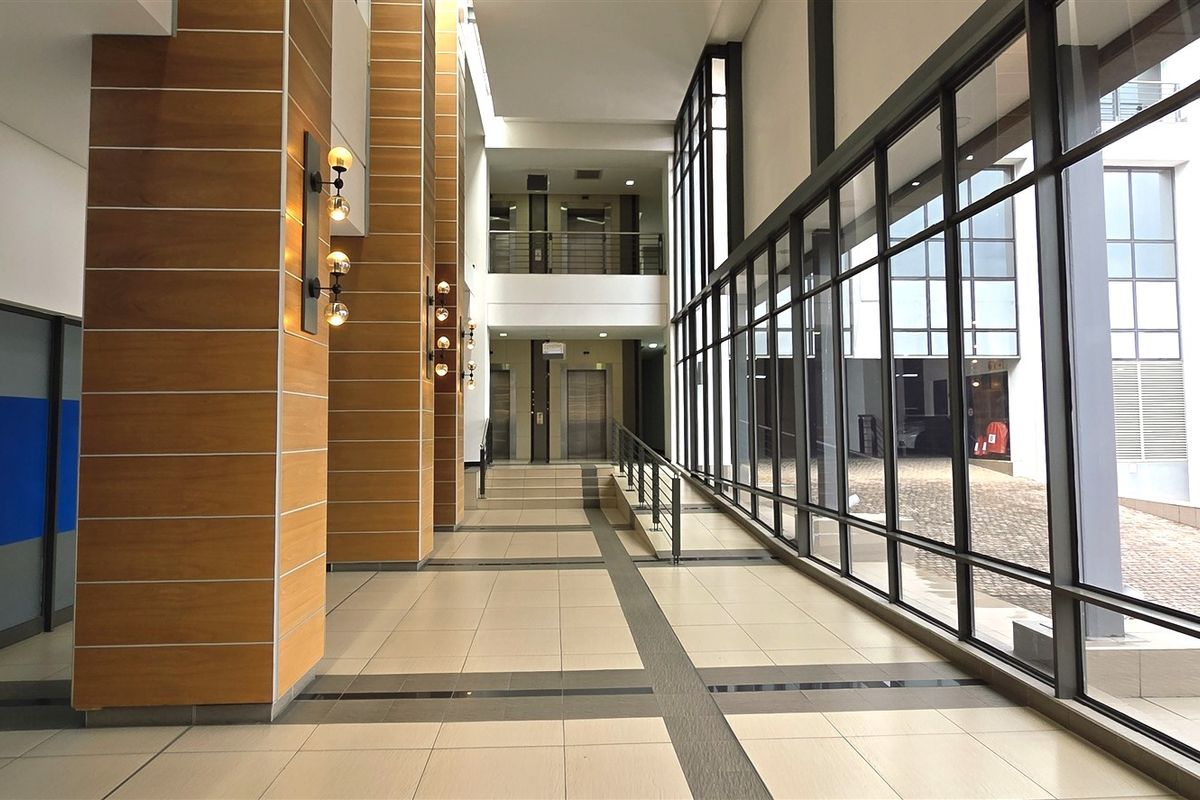 Spacious modern office building lobby featuring floor-to-ceiling glass walls, wood-panelled columns, tiled flooring, and elevator access.