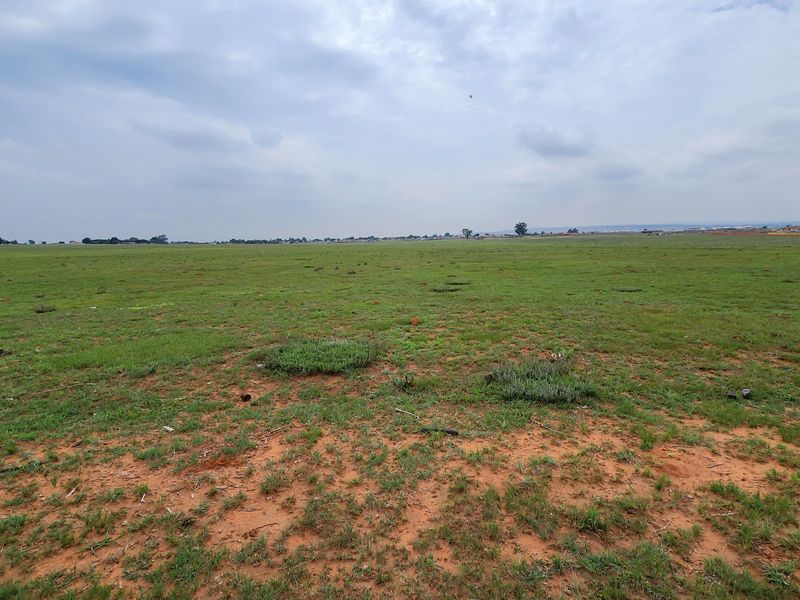 A wide-angle view of a large, flat, undeveloped commercial land plot featuring open grassland under a cloudy sky.