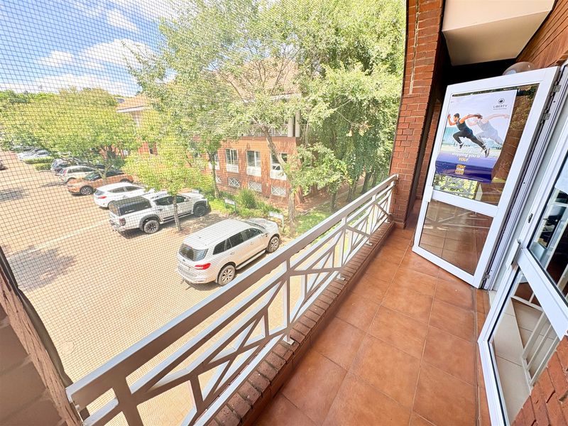 A covered balcony featuring tiled flooring, a brick wall, and a metal balustrade, overlooking the office complex parking area.