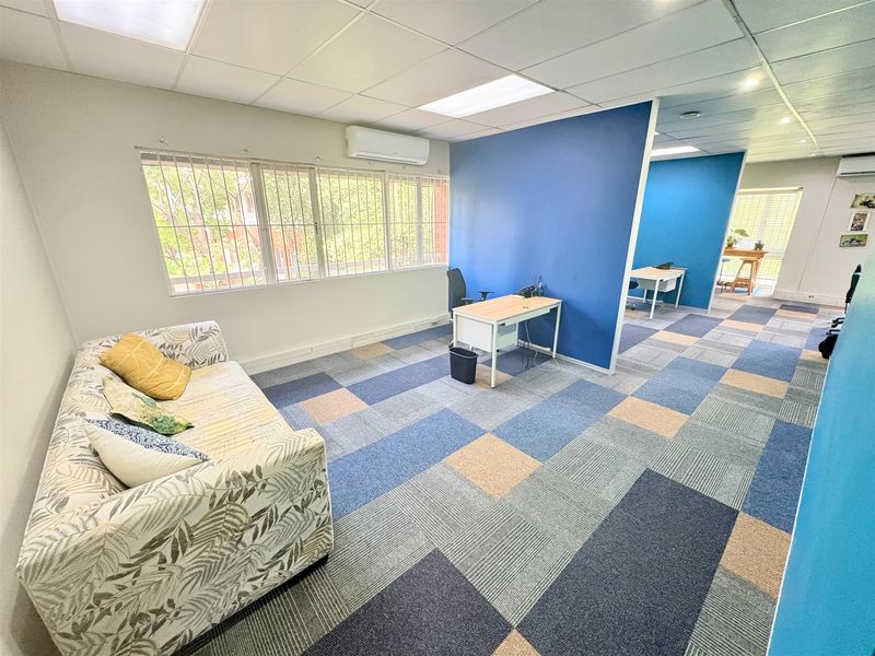 Open-plan office space featuring carpet tiles, suspended ceiling with recessed lighting, split-system air conditioning, and large windows with security bars.