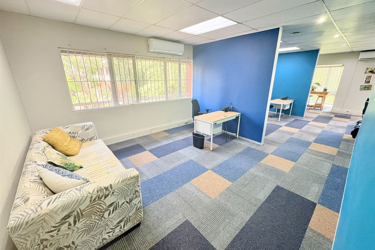 Open-plan office space featuring carpet tiles, suspended ceiling with recessed lighting, split-system air conditioning, and large windows with security bars.