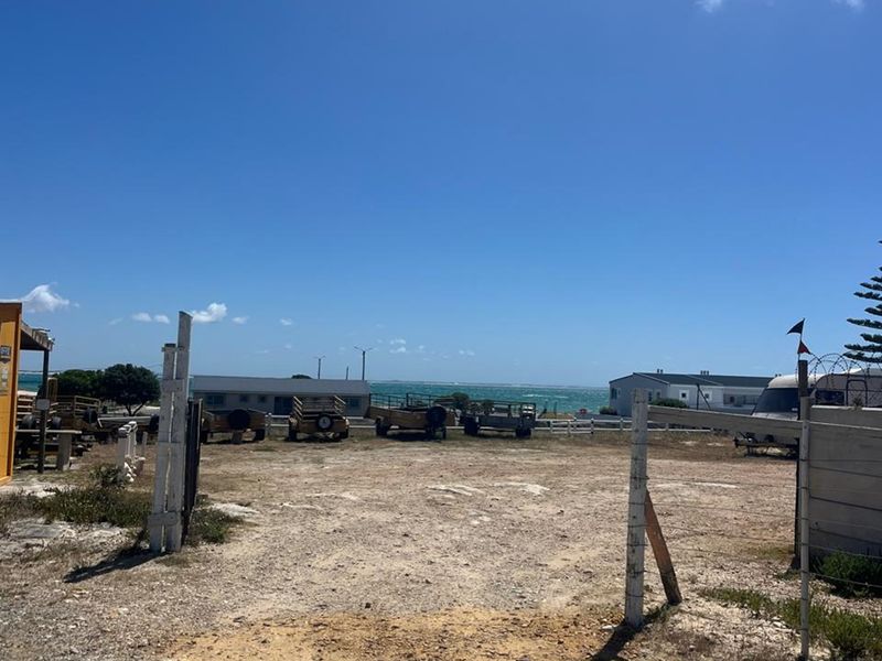 An open, unpaved outdoor area with perimeter fencing, offering a clear view towards the coastline.