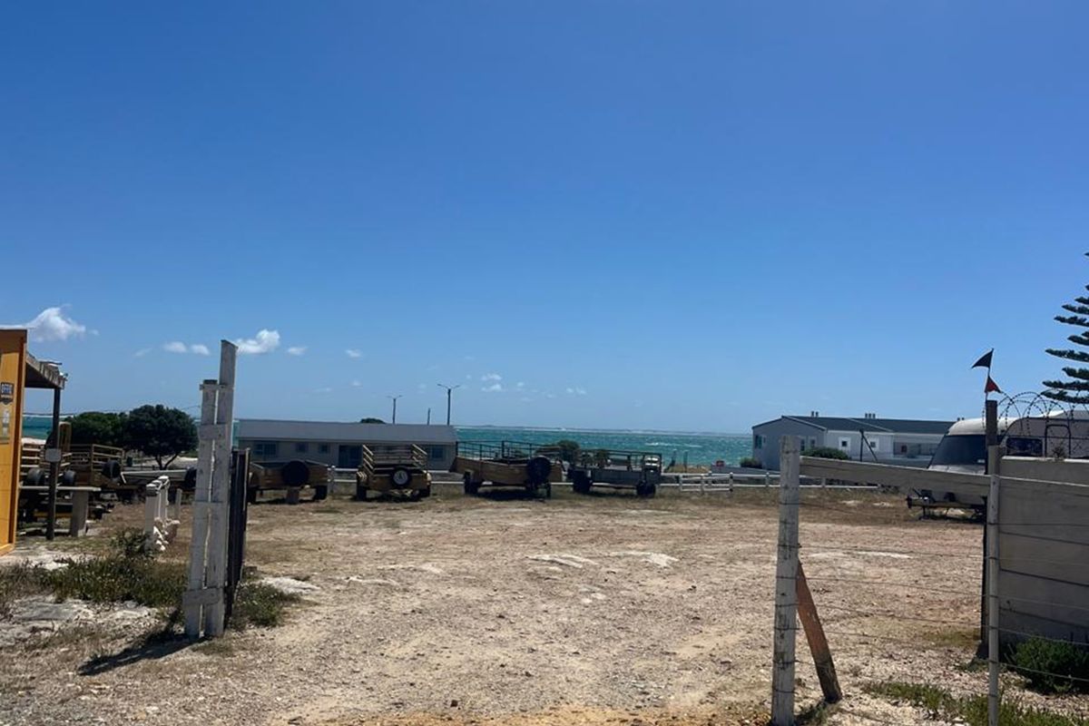 An open, unpaved outdoor area with perimeter fencing, offering a clear view towards the coastline.