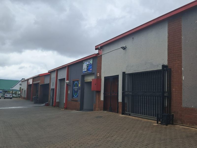 Exterior view of a commercial retail unit complex featuring paved frontage, roller shutter doors, and secure metal gate access.