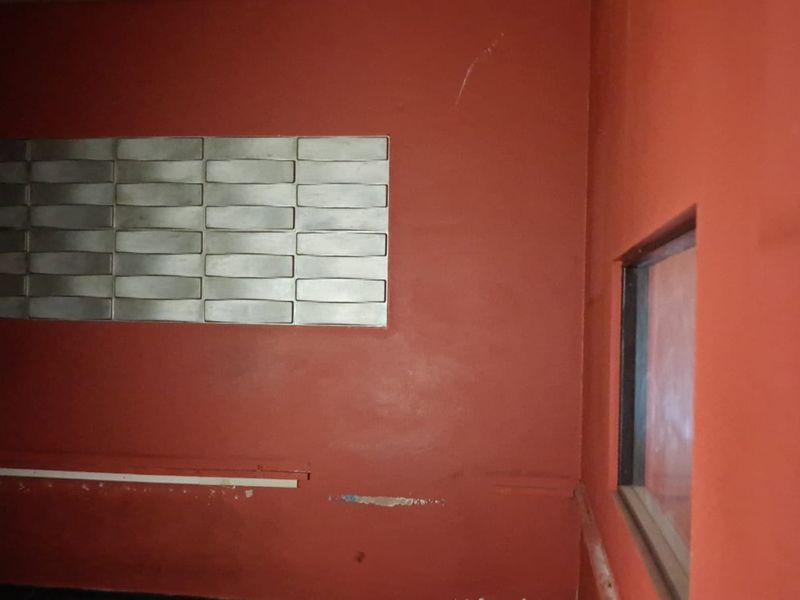 An interior retail space featuring red painted walls, a decorative metallic wall panel, and a side window, currently in need of cosmetic refurbishment.
