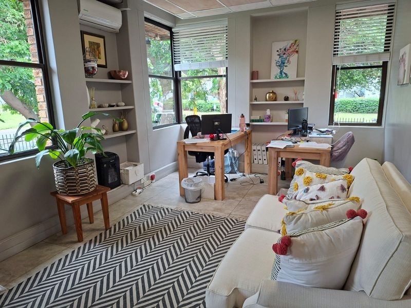 A bright office space featuring built-in shelving, tiled flooring, large windows with blinds, and a wall-mounted air conditioning unit.