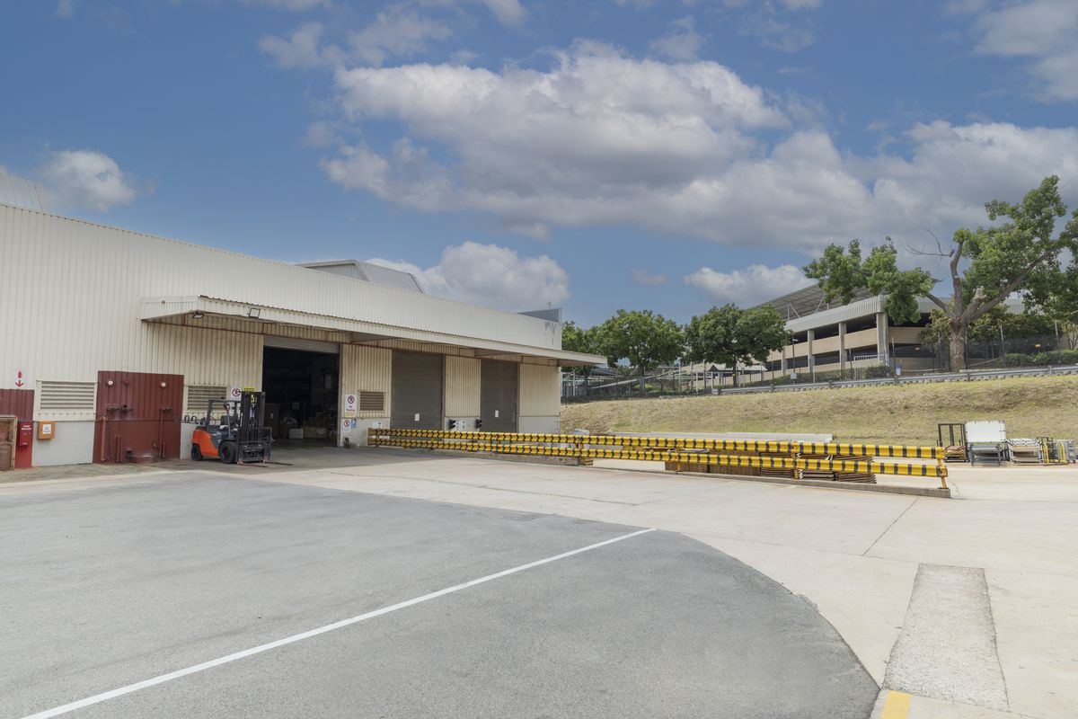 Industrial warehouse exterior featuring a concrete loading yard, roller shutter access doors, and protective safety barriers.