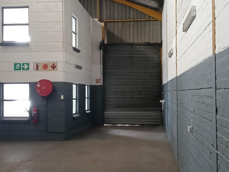 Industrial warehouse space featuring high ceilings, exposed steel beams, concrete flooring, and a large roller shutter door for vehicle access.