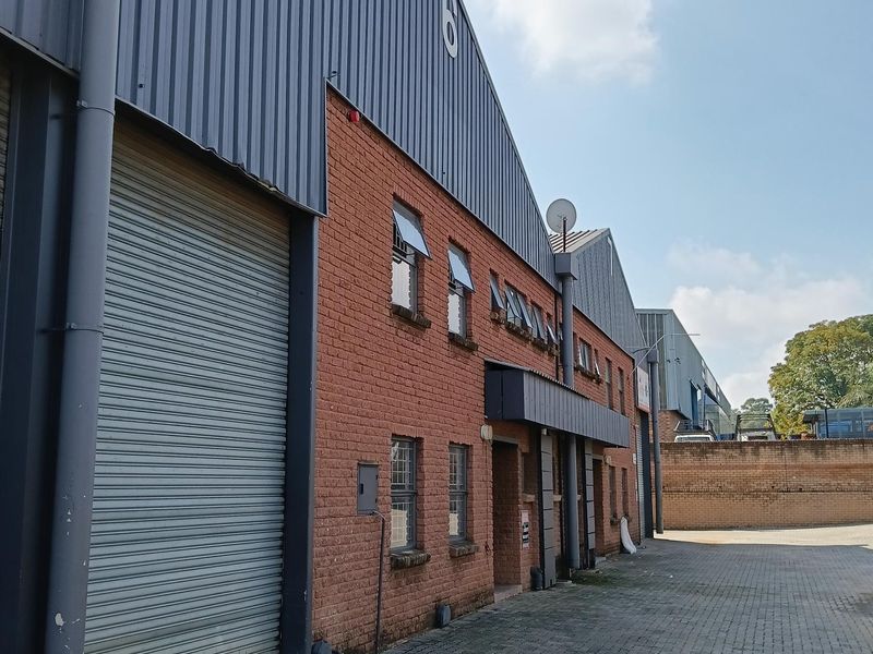 Exterior view of an industrial warehouse unit featuring brick facade, metal cladding, and a large roller shutter door with paved access area.
