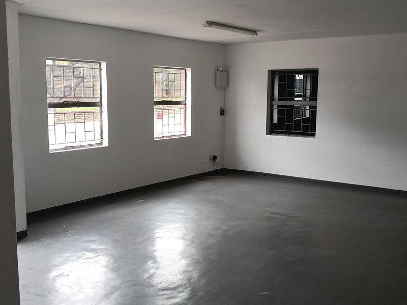 An empty industrial office space featuring painted concrete flooring, white walls, and security-barred windows providing natural light.