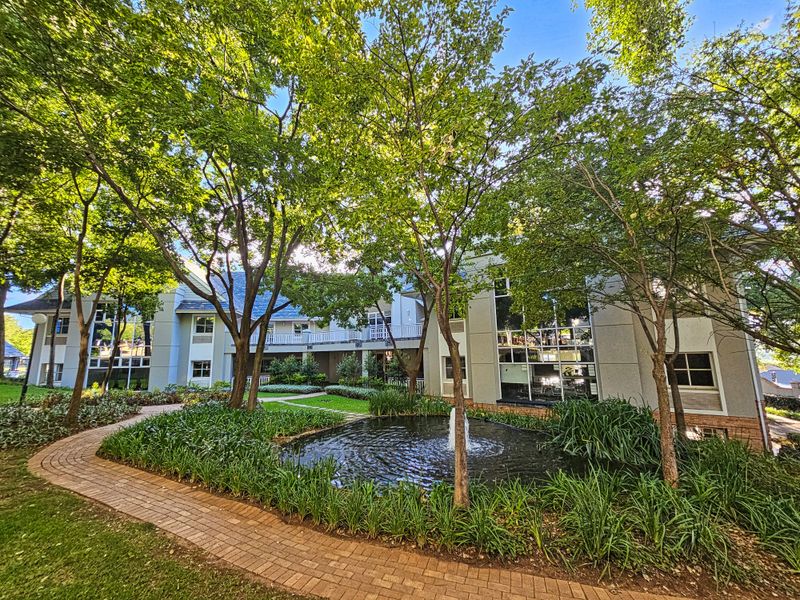 A well-maintained office building exterior featuring a landscaped garden, a central water feature, and a brick-paved pathway leading to the building entrance.