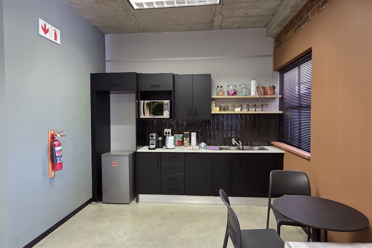 A compact office kitchenette featuring modern black cabinetry, a stainless steel sink, tiled backsplash, and an industrial-style exposed concrete ceiling.