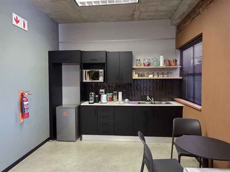 A compact office kitchenette featuring modern black cabinetry, a stainless steel sink, tiled backsplash, and an industrial-style exposed concrete ceiling.