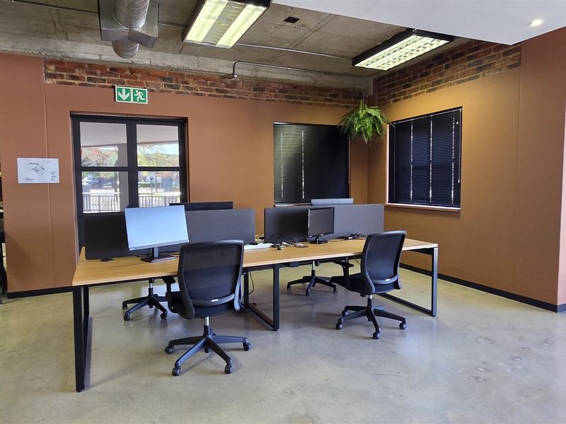 Modern open-plan office space featuring exposed concrete ceilings, brick accent walls, polished concrete flooring, and an integrated kitchenette area.