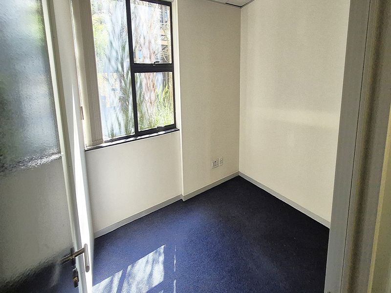 A compact, empty office space featuring blue carpet flooring, a suspended ceiling, and a window providing natural light.
