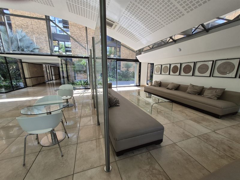 Modern office lounge area featuring high ceilings, large glass partitions, tiled flooring, and extensive natural light through floor-to-ceiling windows.