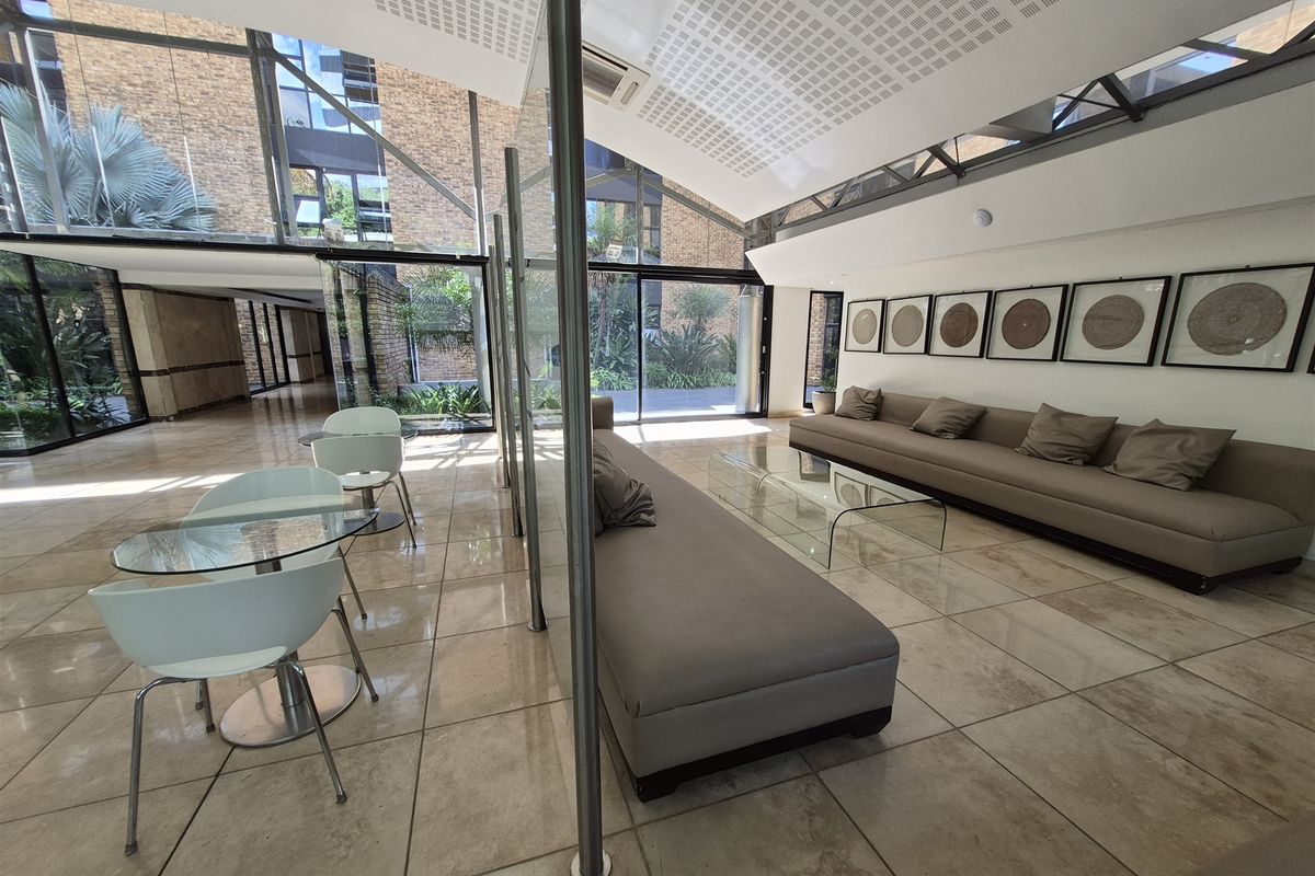 Modern office lounge area featuring high ceilings, large glass partitions, tiled flooring, and extensive natural light through floor-to-ceiling windows.