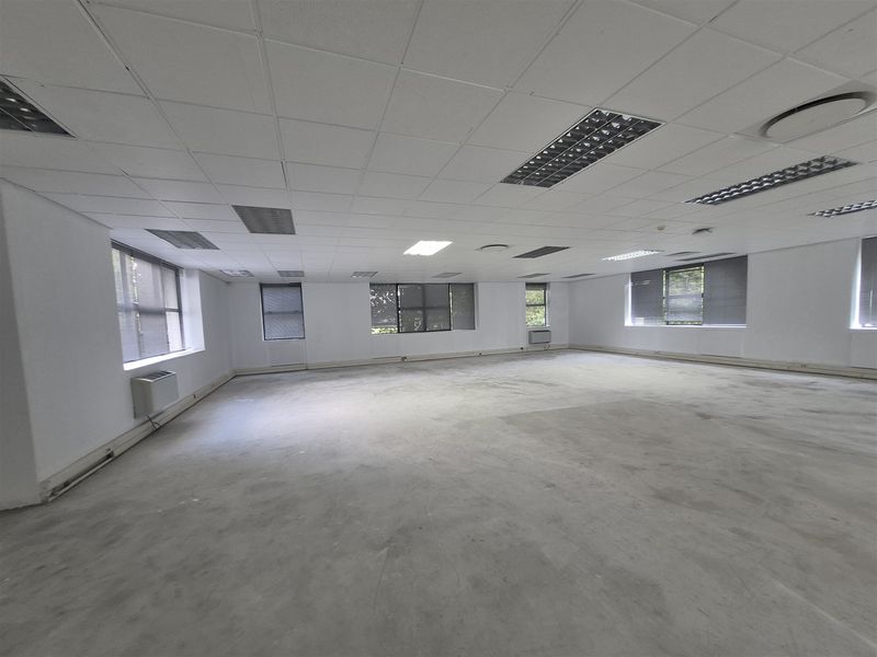 A spacious, vacant open-plan office area featuring a suspended ceiling, recessed lighting, air conditioning units, and large windows with fitted blinds.