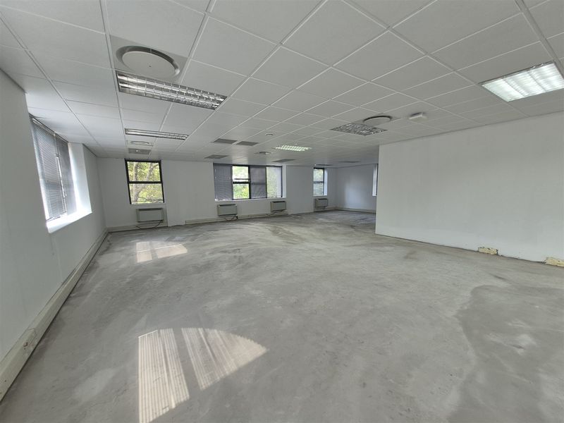 An expansive, empty office space featuring a suspended ceiling, recessed lighting, integrated air conditioning units, and large windows with fitted blinds.