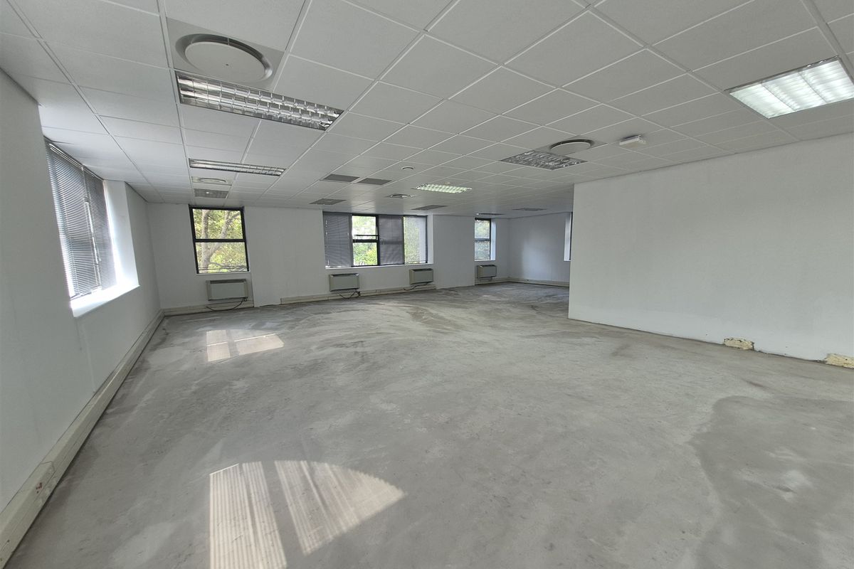 An expansive, empty office space featuring a suspended ceiling, recessed lighting, integrated air conditioning units, and large windows with fitted blinds.