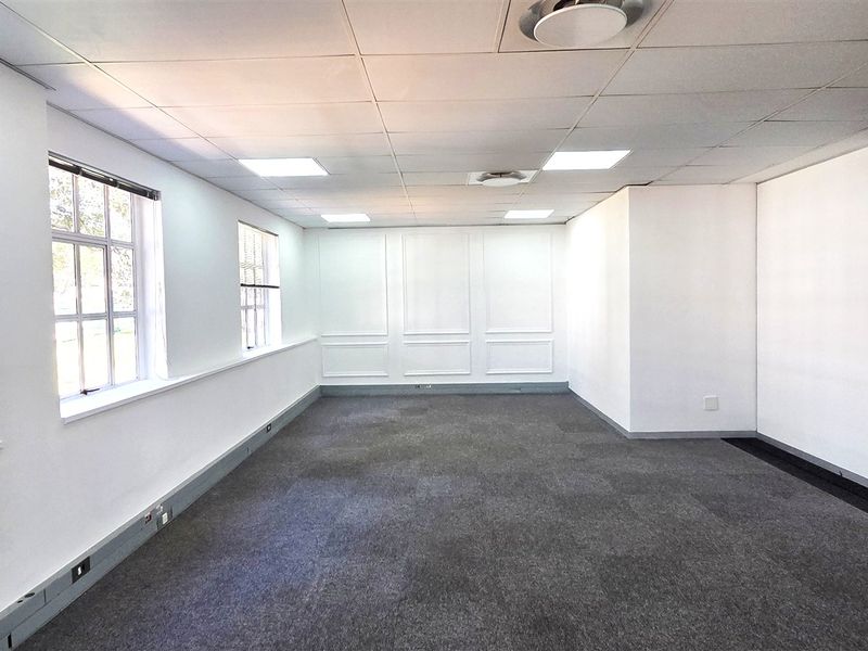 A bright, empty office space featuring a suspended ceiling, recessed lighting, carpeted flooring, and elegant wainscoting, with access via a sliding glass door.