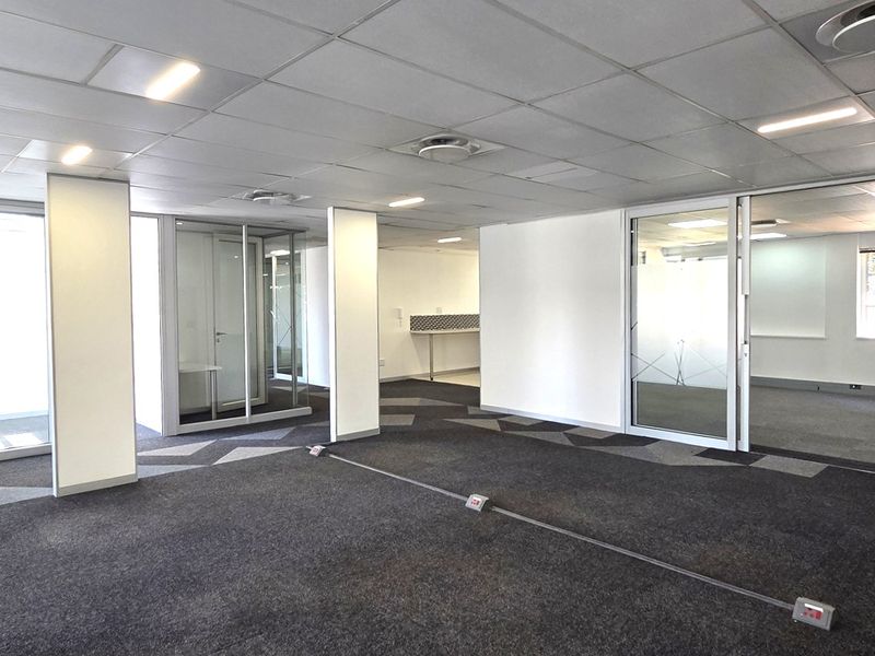 A spacious, open-plan office area featuring suspended ceilings, recessed lighting, glass partitions, and carpeted flooring, offering a clean and professional workspace environment.