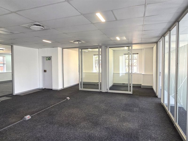 A bright, open-plan office space featuring modern glass partitions, suspended ceilings with recessed lighting, and clean carpet flooring.