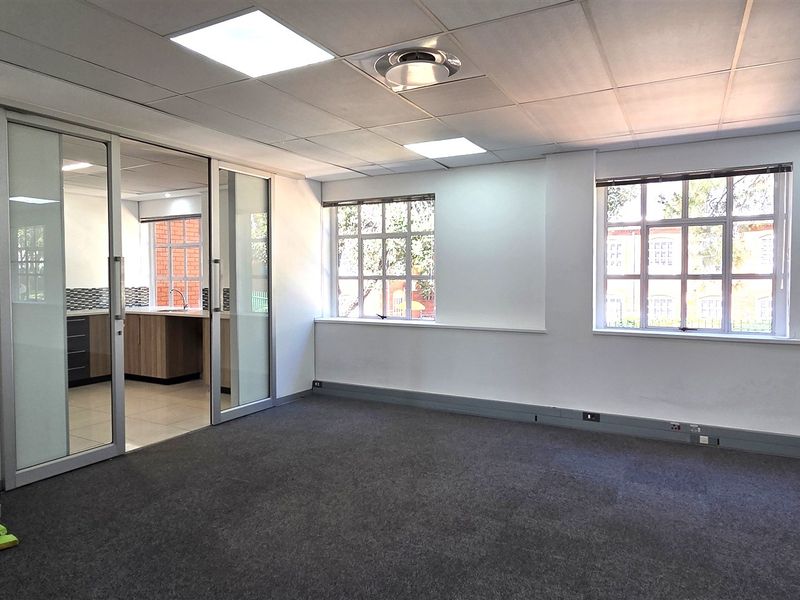 A bright, open-plan office space featuring carpeted flooring, a suspended ceiling with recessed lighting, and a sliding glass partition leading to a kitchenette area.